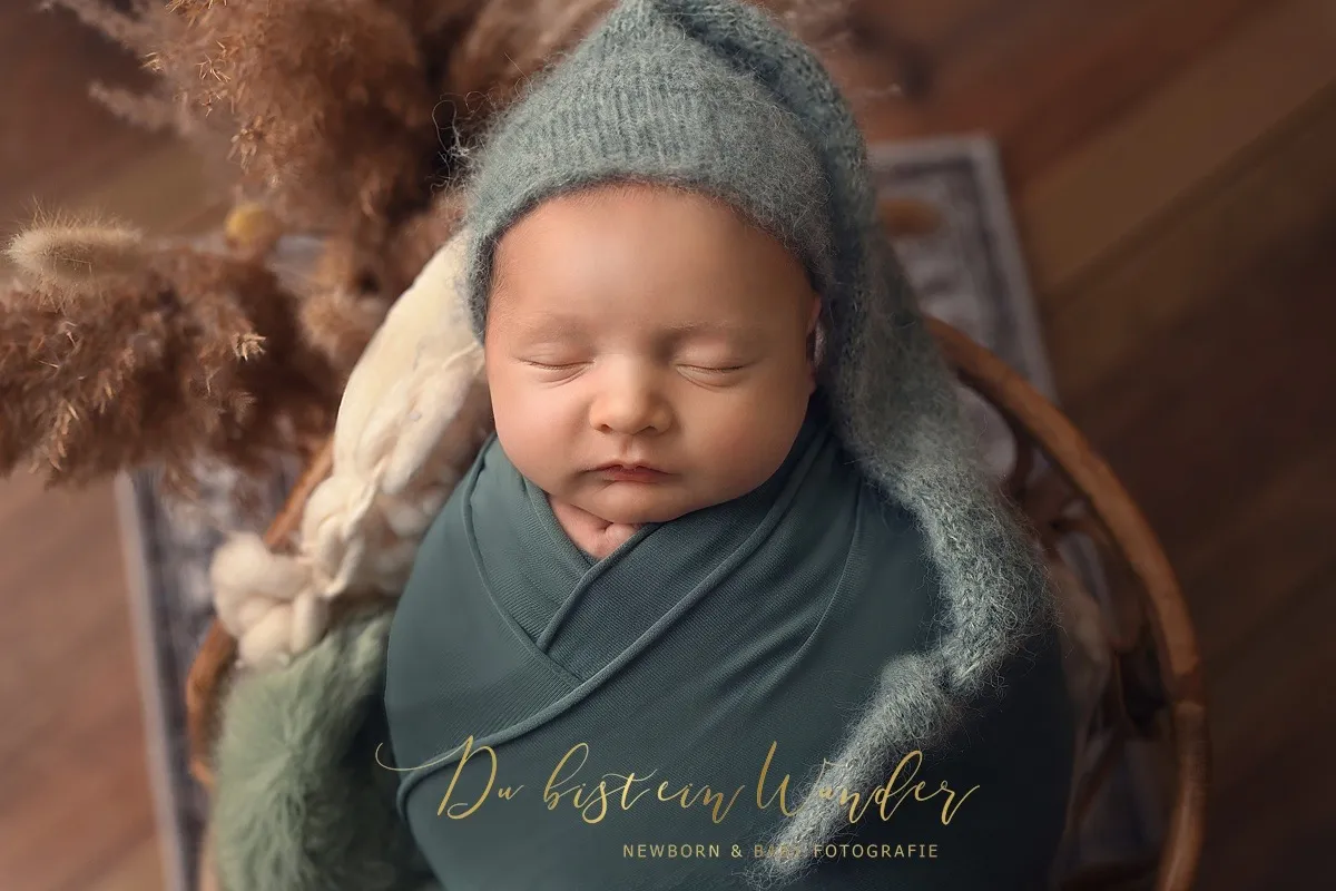 Schlafendes Baby mit grüner Strickmütze und Wrap im Weidenkorb mit Pampasgras