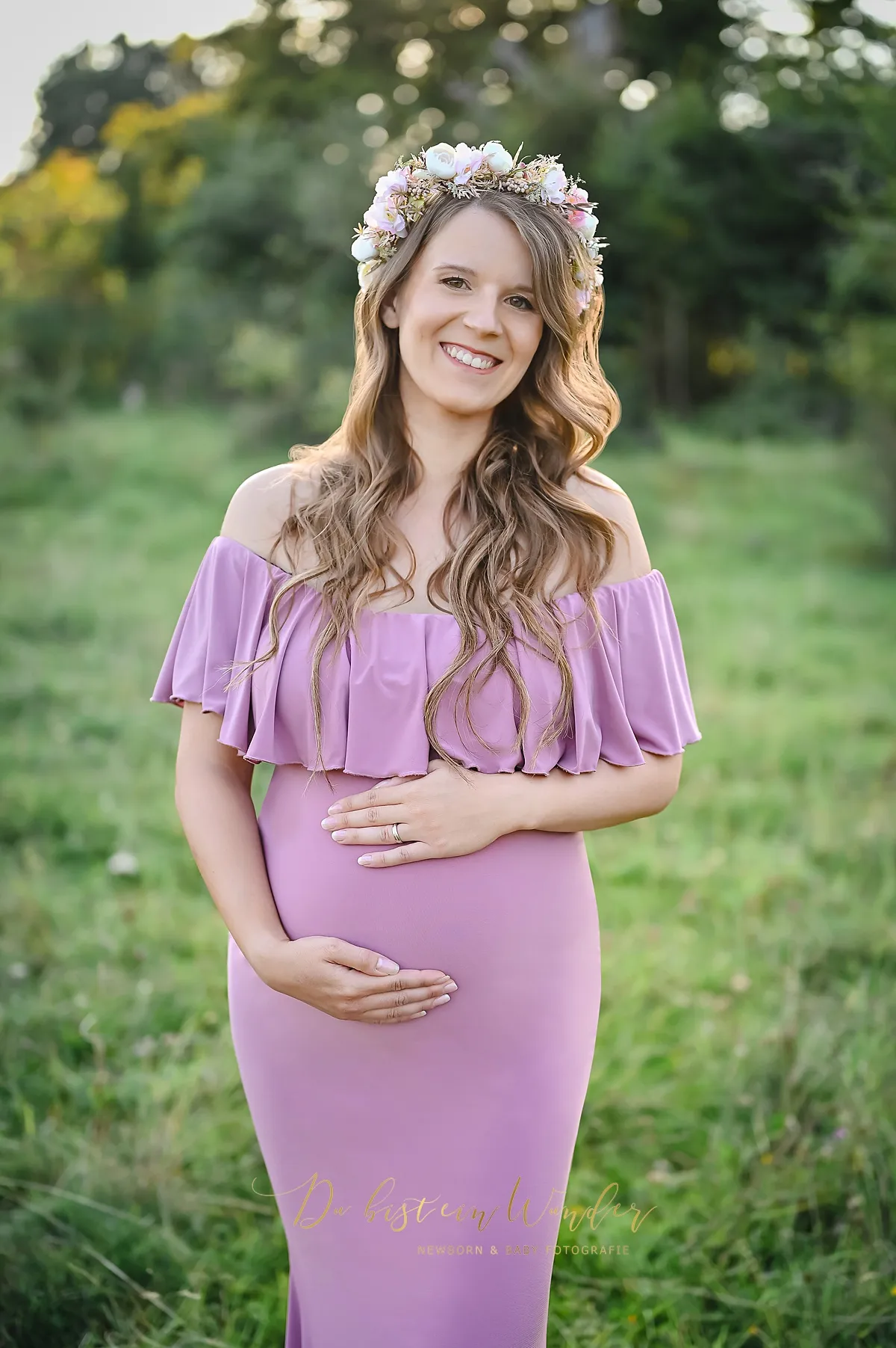 Lächelnde Schwangere auf der Wiese im rosa Kleid mit Blumenkranz