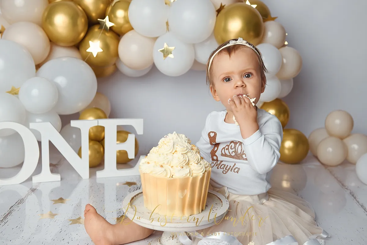 Kleines Mädchen nascht vom Kuchen beim Cake-Smash Shooting mit goldener Ballondeko