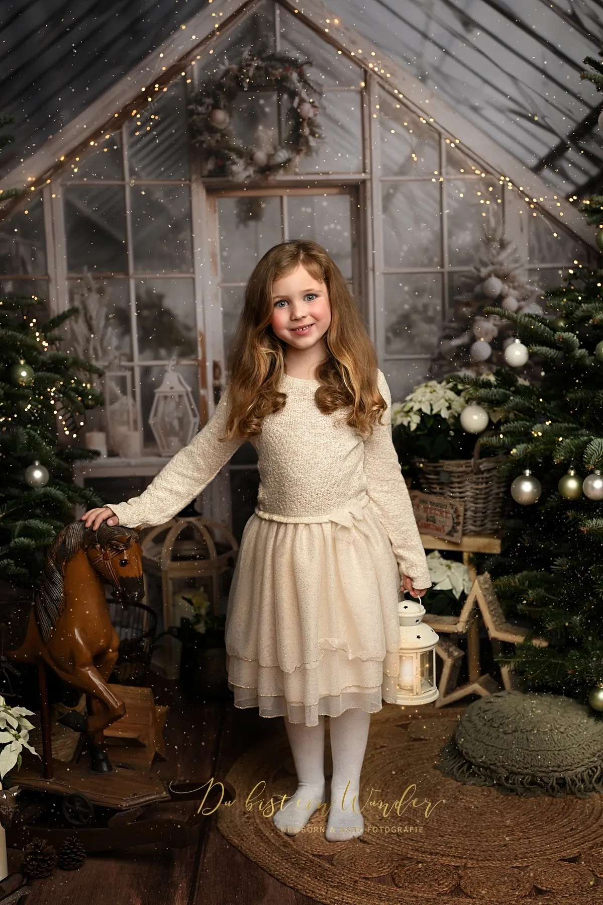 Mädchen im weißen Kleid steht im weihnachtlich geschmückten Wintergarten mit Laterne und Schaukelpferd