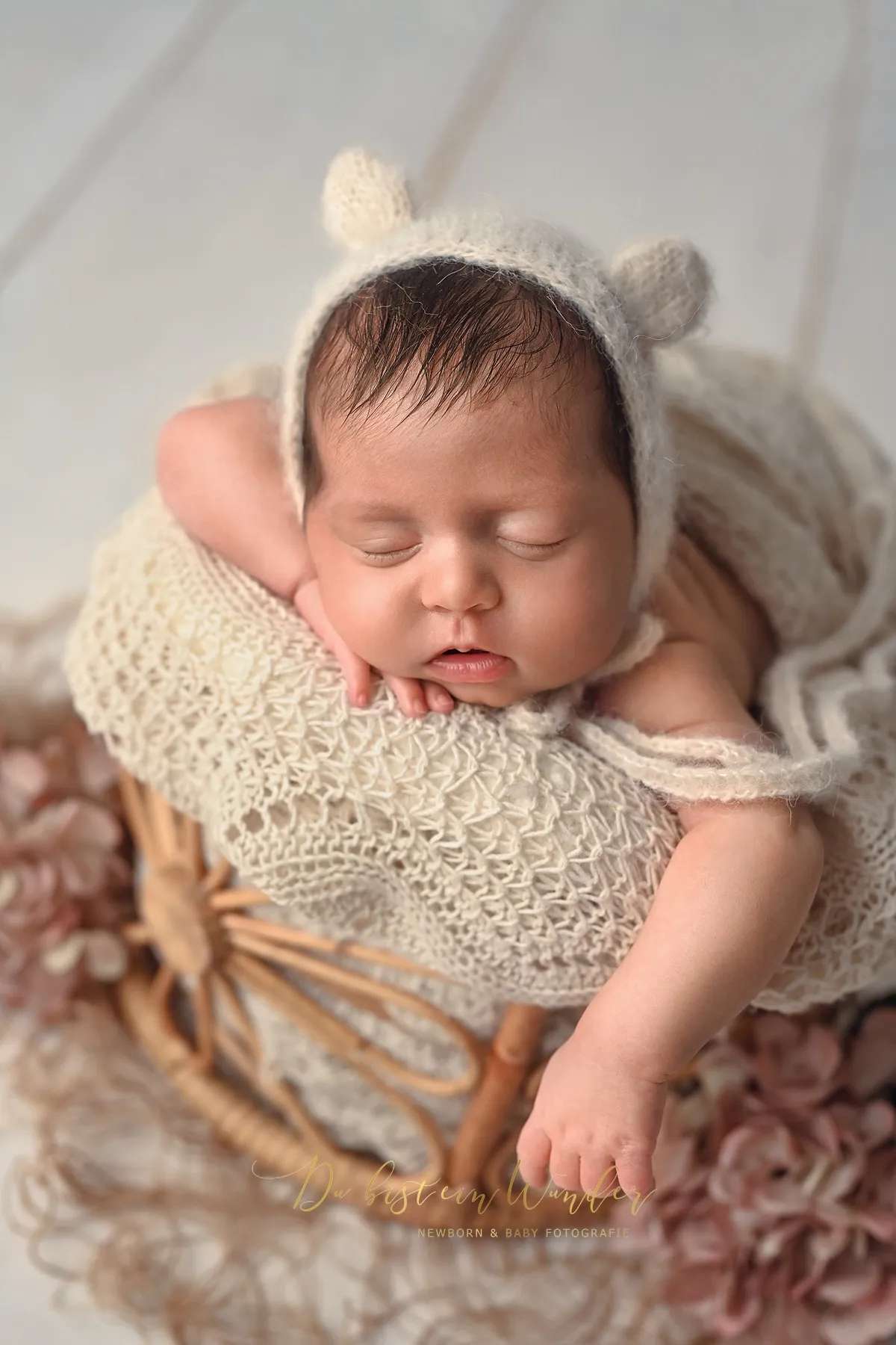 Schlafendes Neugeborenes mit gestrickter Bärenohr-Haube in einem Körbchen mit rosa Blumen