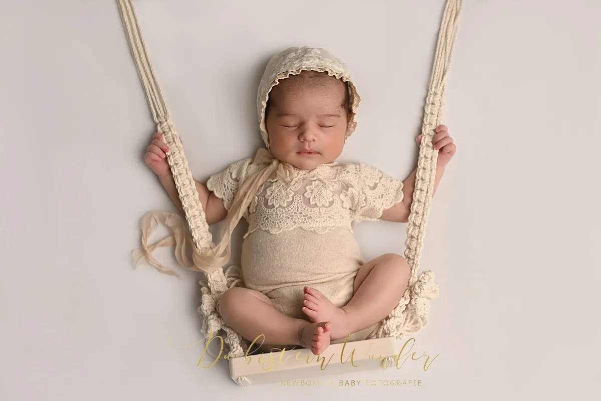 Schlafendes Neugeborenes im Spitzenoutfit und Häubchen auf einer Makramee-Schaukel