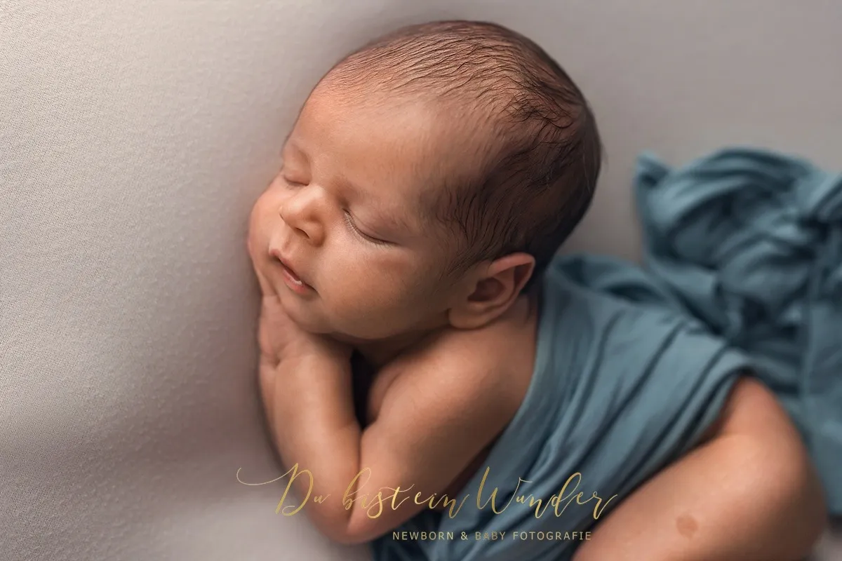 Nahaufnahme eines schlafenden Neugeborenen in blauem Tuch mit gefalteten Händen